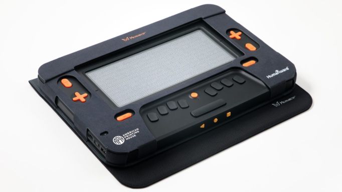 Photo of a Monarch 2D display.  It is black, about the size of a typical book, and has large raised pale orange buttons for controls. It also has ports for connectivity on the edges.